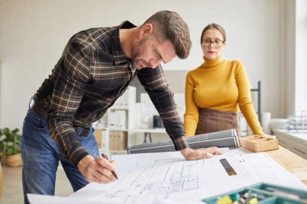 Side view portrait of mature bearded architect drawing blueprints while standing by desk in office with female colleague in background
