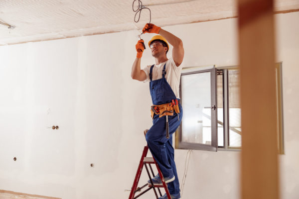 Handsome young man in safety helmet standing on ladder and using pliers while repairing electrical wiring